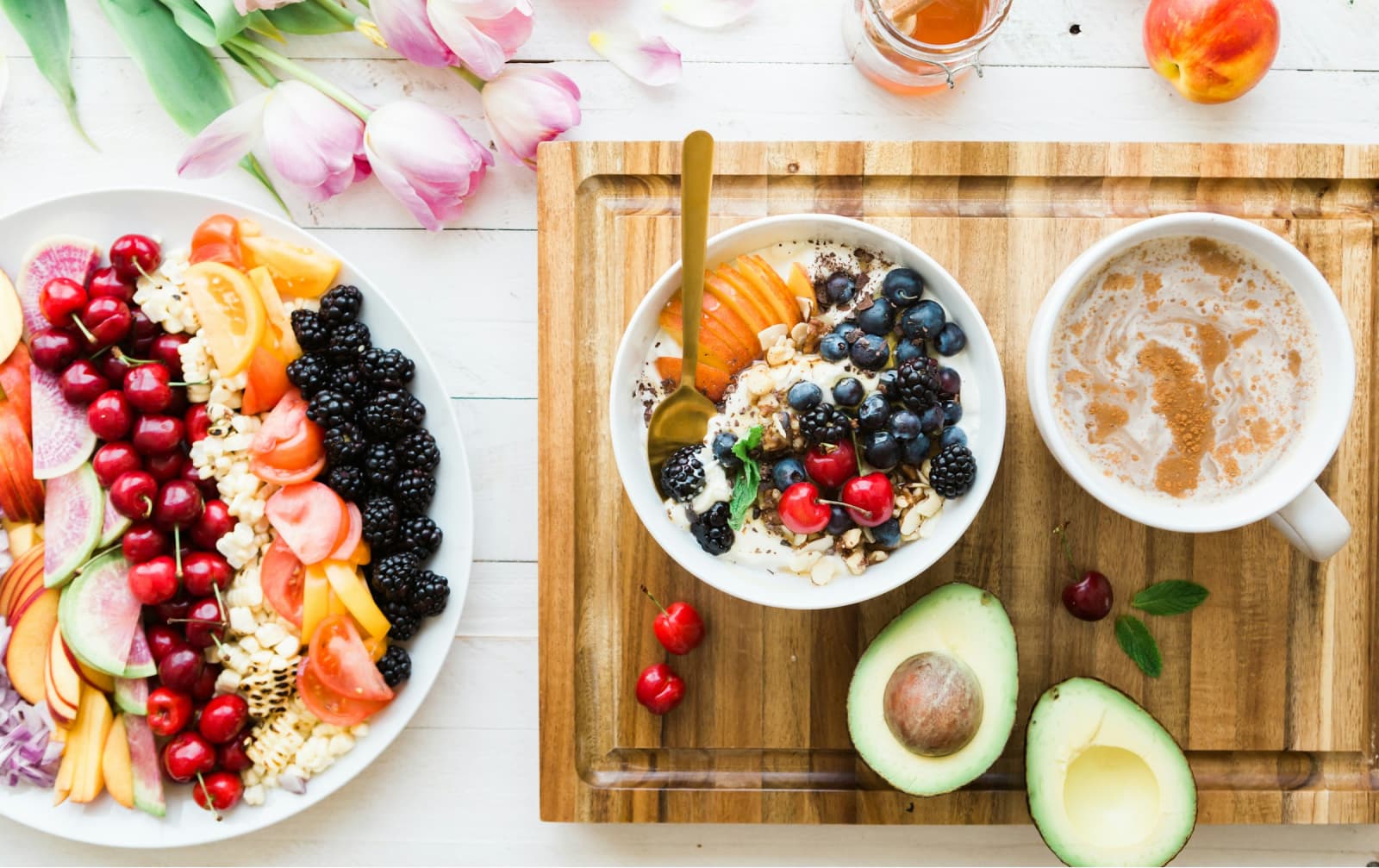 A wood board with an avocado, a coffee, a yogurth bowl with fruit and a salad fruit to the side, some flowers in the left top corner and some honey