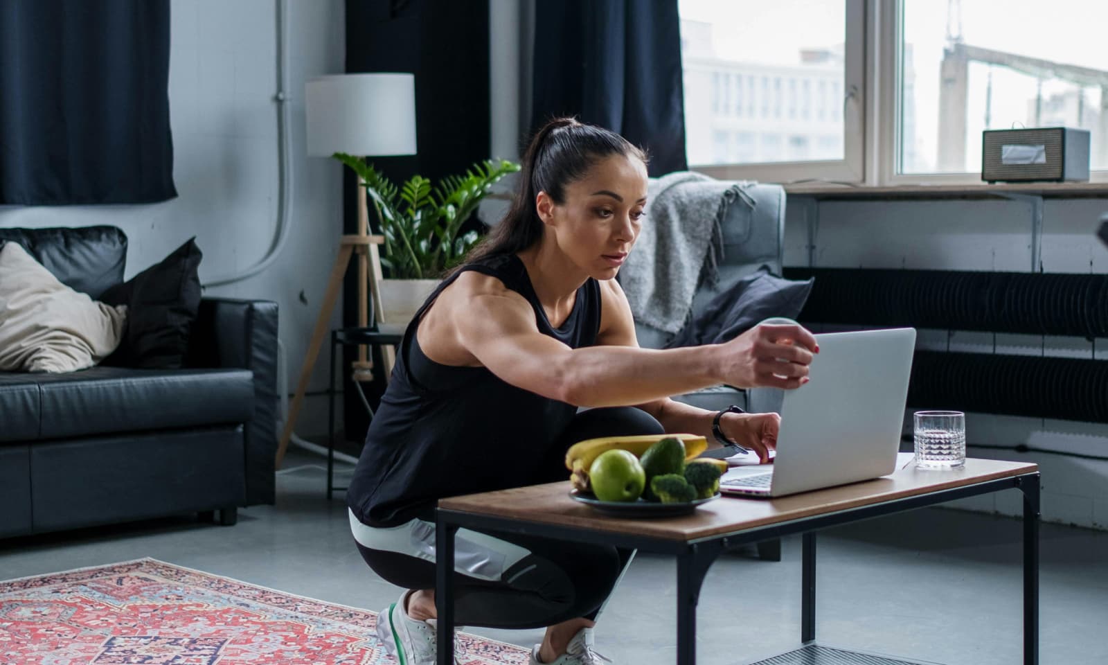 Nutrition coach crouching in front of a table with a laptop, fruits and a glass of water in a modern apartment